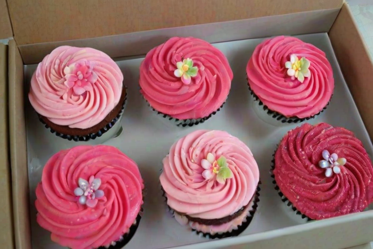 Create an image of a box containing a dozen beautifully decorated cupcakes. The cupcakes should feature various shades of pink frosting, some with intricate designs, flower decorations, and edible glitter. Include a mix of smooth swirls, piped rosettes, and decorative toppings like sprinkles and small fruit pieces. The box should be open, showcasing the delightful array of cupcakes inside.