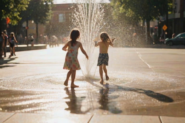 Blurry, candid photo of children playing in a fountain on a hot summer day, water spraying in all directions, with distant buildings casting long shadows on the street.