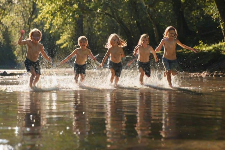 Blurry action shot of a group of kids splashing in a shallow creek, water droplets suspended in mid-air, sunlight reflecting off the water, the distant sound of laughter and rushing water, soft shadows cast by trees on the banks.