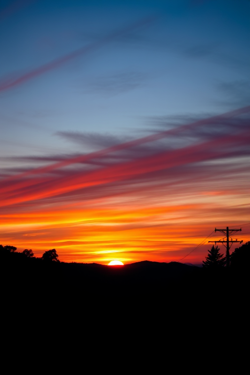Capture a vibrant sunset over hills with silhouettes of trees and utility poles, using a DSLR camera, aperture f/8, shutter speed 1/200 sec, ISO 100, wide-angle lens, and a low angle view to enhance color saturation and contrast.