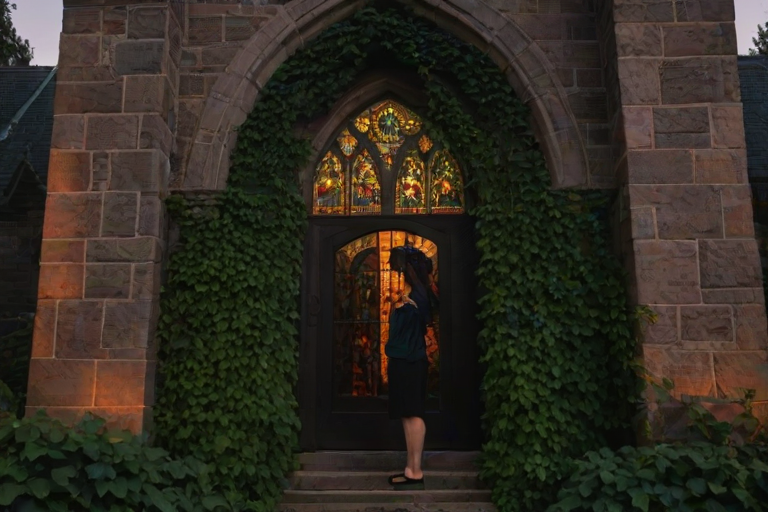 Old stone church at twilight, with ivy climbing its walls, a woman stands in front of the entrance, her silhouette framed by the last light of day as she gazes up at the stained-glass windows.