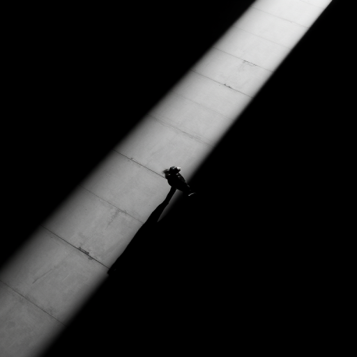 create a high-contrast black and white image of a lone figure standing in a narrow beam of light that cuts through a dark space, with deep shadows and dramatic angles, shot with a 35mm lens at f/1.8, ISO 100, in a minimalistic style using a mirrorless camera from a bird's eye view to emphasize depth and solitude