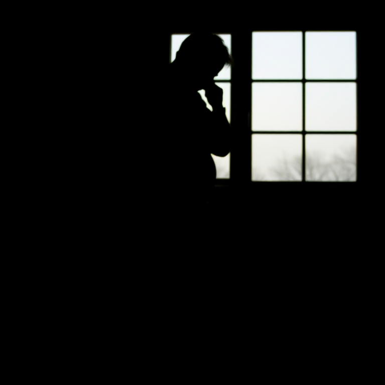 Create a dimly lit image of a person in silhouette with a window in the background, shot with a DSLR camera at f/1.8, ISO 800, shutter speed 1/60s, in portrait view, using a low-angle shot to emphasize the shadow on the floor.