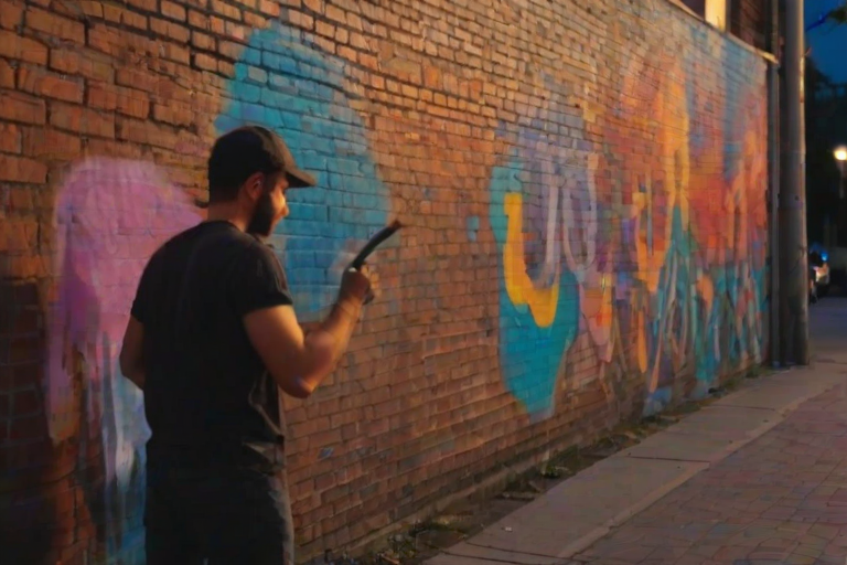 Blurry candid shot of a street artist spray-painting a mural on a brick wall at dusk, vibrant colors blending together, with distant passersby watching and warm light from street lamps casting shadows.