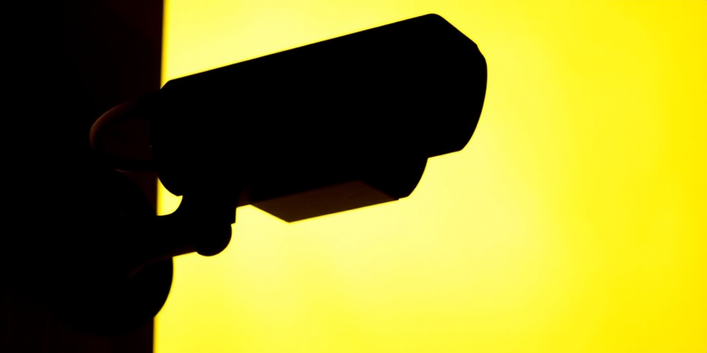 Create a high-resolution silhouette image of a security camera on a bright yellow background, using a DSLR camera with a 50mm lens, f/2.8 aperture, ISO 100, shot from a slightly overhead angle to improve depth, focus, and clarity.