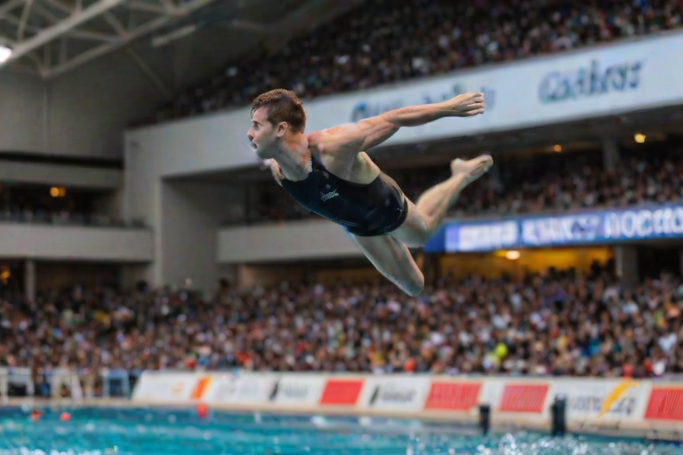 Blurry action shot of a diver leaping off a high platform, the water below rippling, distant spectators watching from the stands, and the intense focus on the grace and speed of the motion.