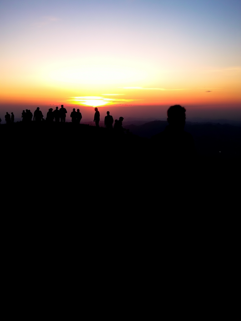 Generate a silhouetted scene of people walking in a line on a hill during sunset, with warm colors blending in the sky, taken with a DSLR camera at f/8, ISO 200, 50mm lens, and a shallow depth of field for clear foreground against a blurred background.