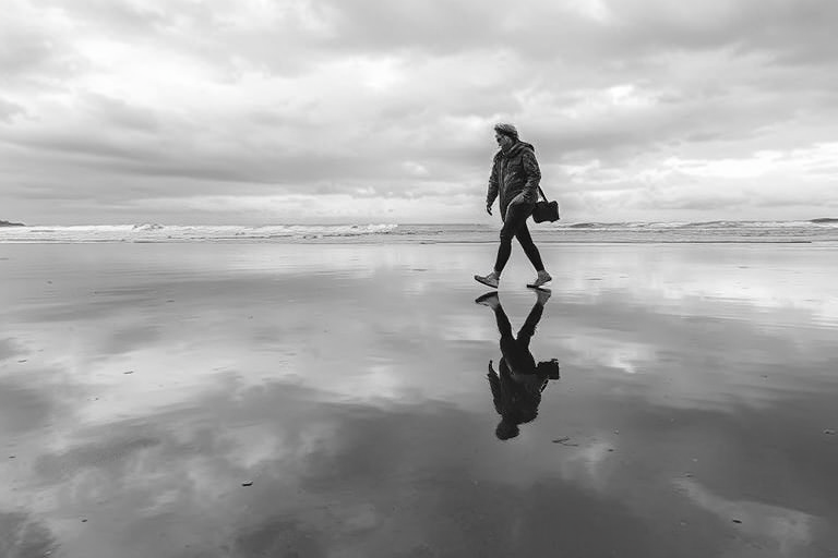 generate a high-contrast black and white image of two people walking on a wet beach during a cloudy day, capturing their reflections on the water, using a full-frame camera with a 50mm lens, aperture set to f/1.8, shutter speed at 1/500s, ISO 100, shot from a low angle to emphasize the reflections and the horizon in the background
