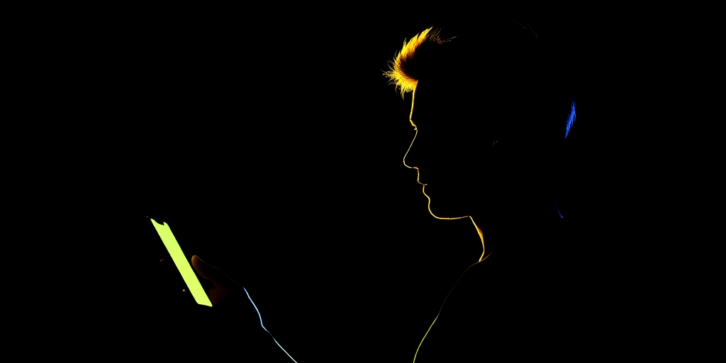 Create a high-contrast image of a silhouette of a person holding a phone, wearing earbuds, with the outline in bright blue and yellow against a solid black background, using a DSLR camera with a 50mm lens, aperture f/1.8, ISO 100, and focused on the subject in profile.