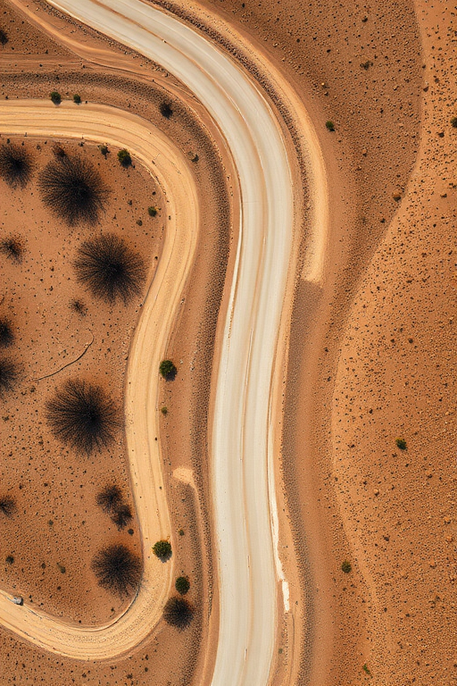 Aerial view of a winding dirt road surrounded by arid landscape, captured with a DSLR camera at 24mm, f/8 aperture, ISO 100, using a drone for a top-down perspective to enhance details and clarity.