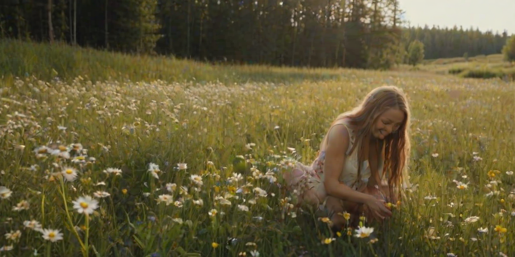 Sunny meadow filled with tall wildflowers, a woman with long braided hair kneels to pick daisies, her laughter echoing softly in the quiet, serene environment.