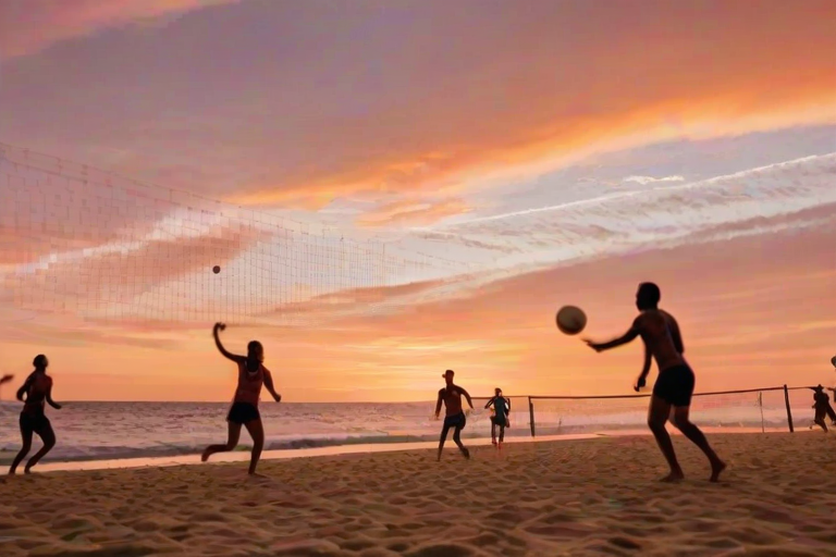 Blurry action shot of a beach volleyball game at sunset, the players silhouettes barely visible against the dramatic orange and pink sky, with soft sand flying as they dive for the ball.