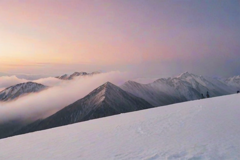 Soft-focus image of a snowy mountain peak under a pastel-colored sunset sky, with soft mist rolling down the slopes and a lone hiker silhouetted on a ridge.