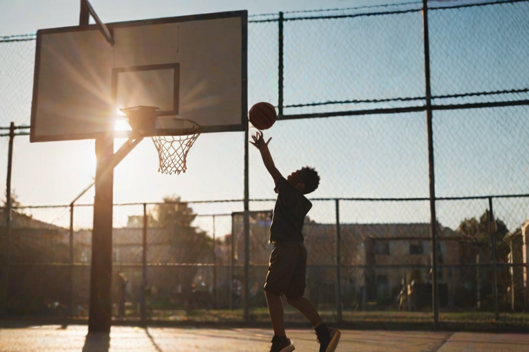 Blurry action shot of a basketball court in an inner city, with a child mid-jump shooting for the basket, and soft sunlight filtering through a chain-link fence in the background.