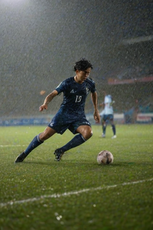 Blurry action shot of a soccer player sliding into a tackle on a rain-soaked field, water droplets flying through the air, with soft stadium lights reflecting off the wet grass and distant teammates watching from the sidelines.