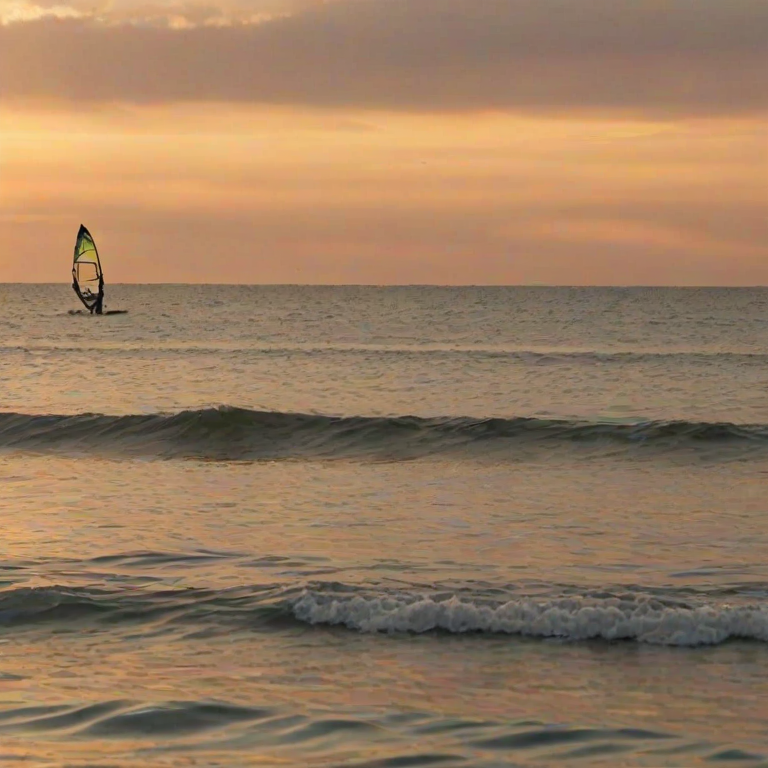Blurry action shot of a windsurfer riding a wave at sunset, soft golden light reflecting off the water, with distant sailboats barely visible on the horizon and a colorful sky overhead.