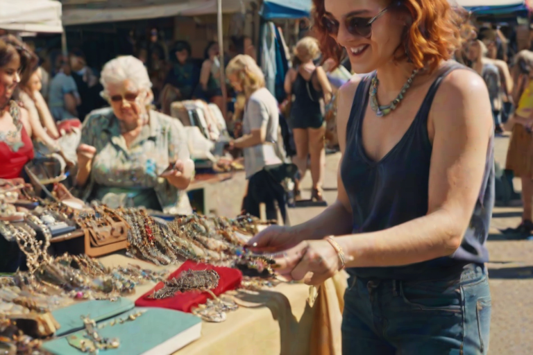 Bustling flea market on a sunny day, a woman browsing through vintage clothes and jewelry, excitedly holding up a unique find to show a friend.