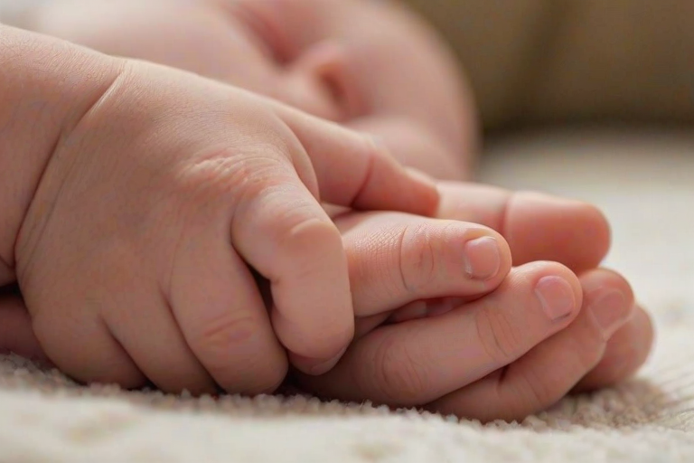 Softly focused close-up of a baby's hand gripping an adult's finger, with soft natural light illuminating their delicate skin, and a cozy blanket in the background providing a sense of warmth and security.