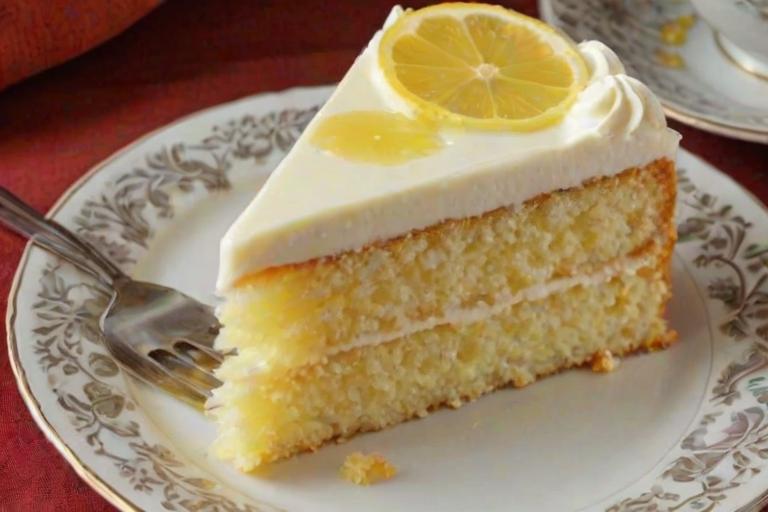 Generate an image of a slice of lemon cake on a decorative plate. The cake should have a light, fluffy texture with a creamy frosting on top, and a lemon filling oozing from the center. Include a lemon slice beside the cake and a golden fork resting on the plate. The background should be soft and neutral to highlight the cake.