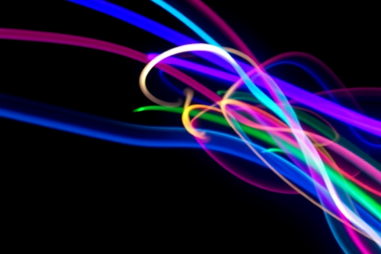 Create an abstract image of colorful light trails on a black background with vibrant blue, pink, green, and yellow hues, using a Canon EOS R5, 50mm lens, f/1.8, ISO 100, and a long exposure of 2 seconds to capture smooth motion and enhance brightness.