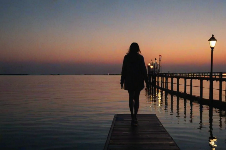 Woman walking along a pier at night, the soft glow of lamplight reflecting off the water, her footsteps echoing as she takes in the peaceful surroundings.