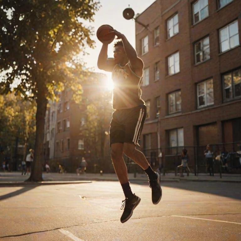 Blurry action shot of a basketball player mid-jump shot in a city playground, with soft golden light from a setting sun casting long shadows on the pavement and nearby buildings.