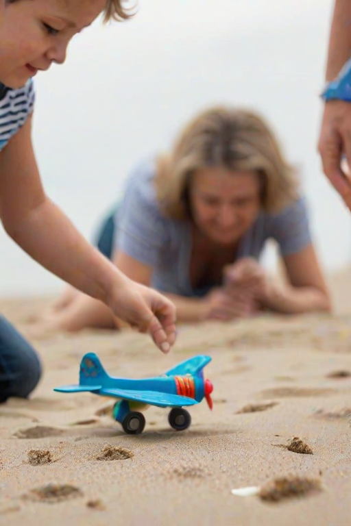 Blurry candid shot of a young boy playing with a toy airplane on a sandy beach, soft waves lapping at the shore, and his mother softly blurred in the background, watching him.